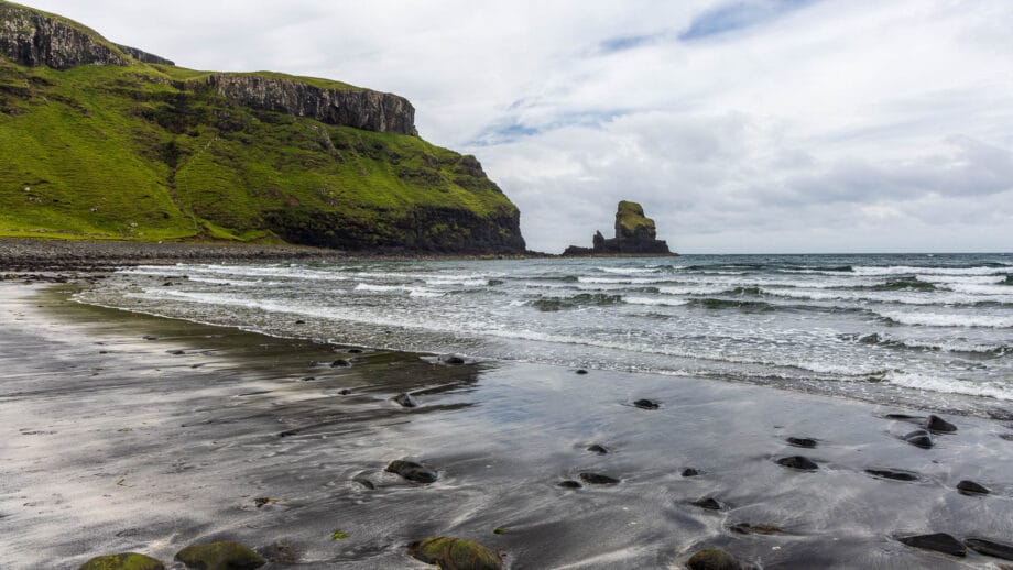 Eine Bucht mit dunklem Sand, einigen Steinen und einer Feldnadel im Hintergrund.