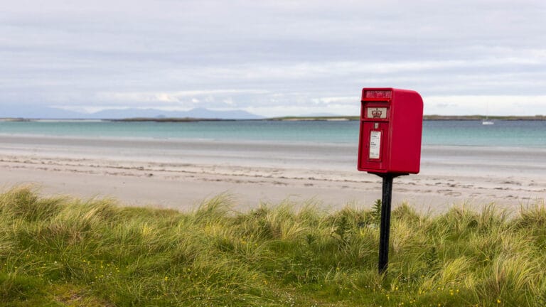 Ein roter Briefkasten der Britischen Post steht vor der Kulisse eines großen weißen Sandstrands.