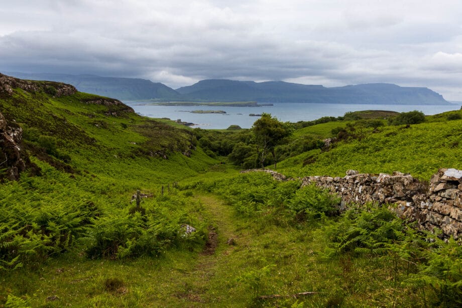 Ein Trampelpfad führt zu einem kleinen Tal hinab, im Hintergrund ist das Meer und die Klippen der Isle of Mull zu erkennen.